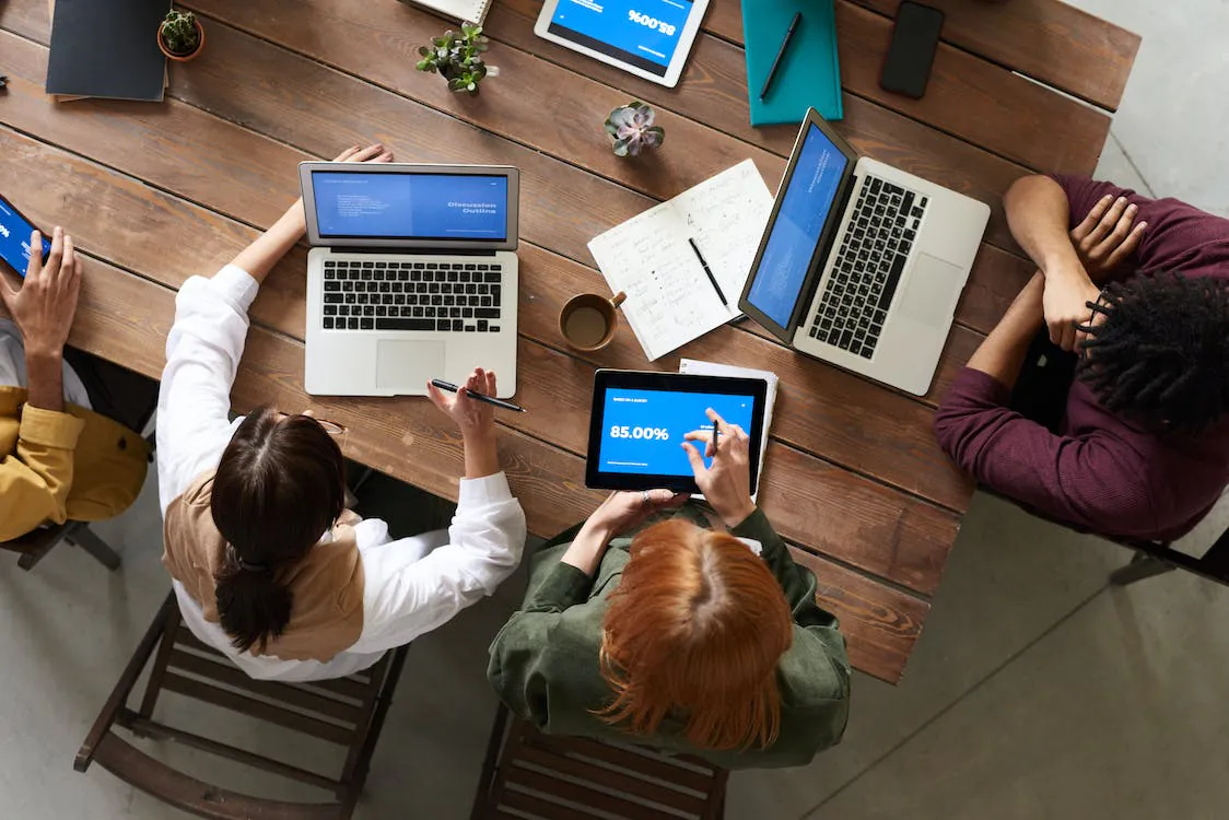 Five individuals sit around a wooden table working on laptops and tablets, with notebooks and coffee cups scattered across the table.