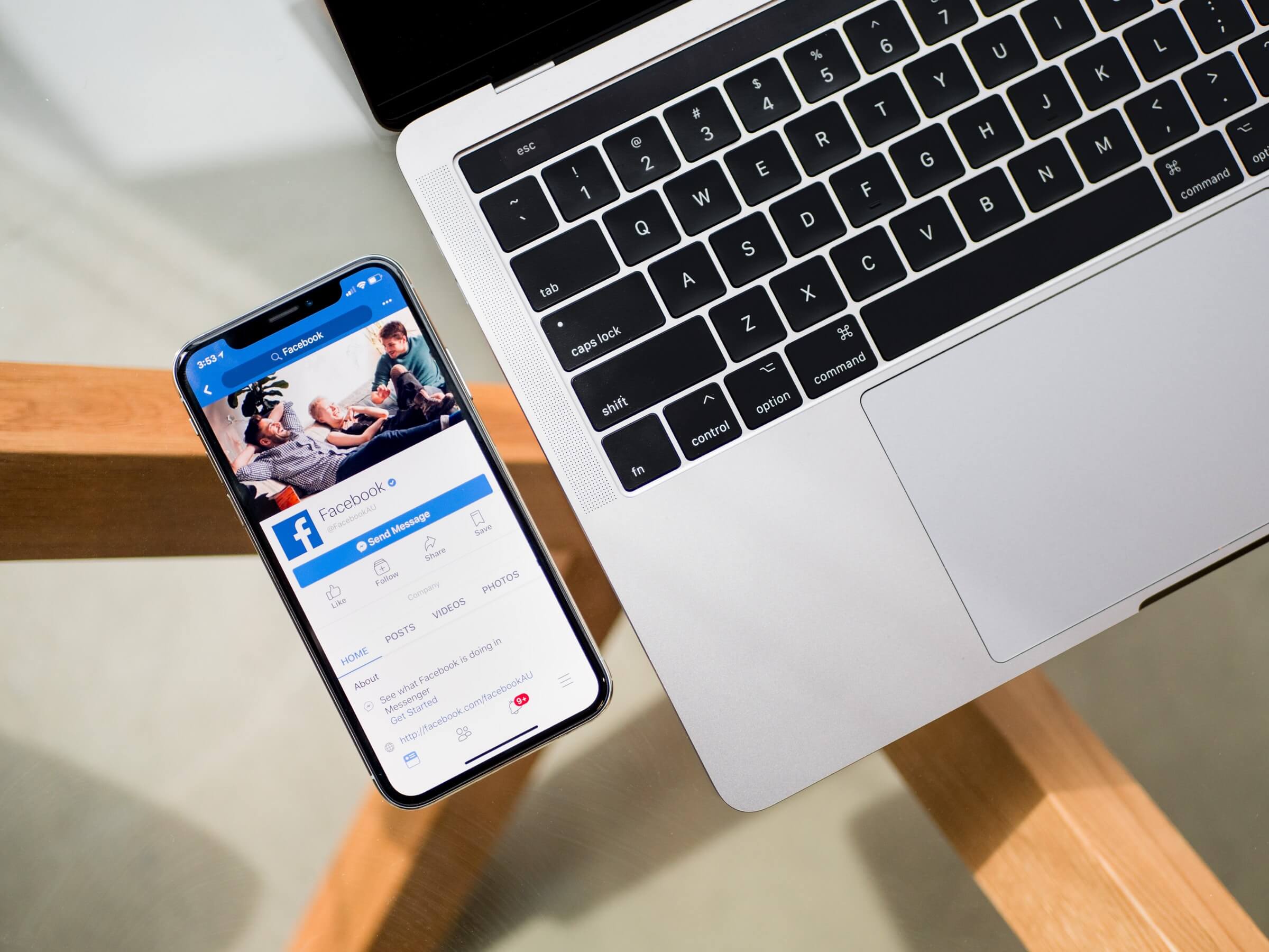 A smartphone displaying a Facebook profile page is placed next to a laptop keyboard on a glass table.