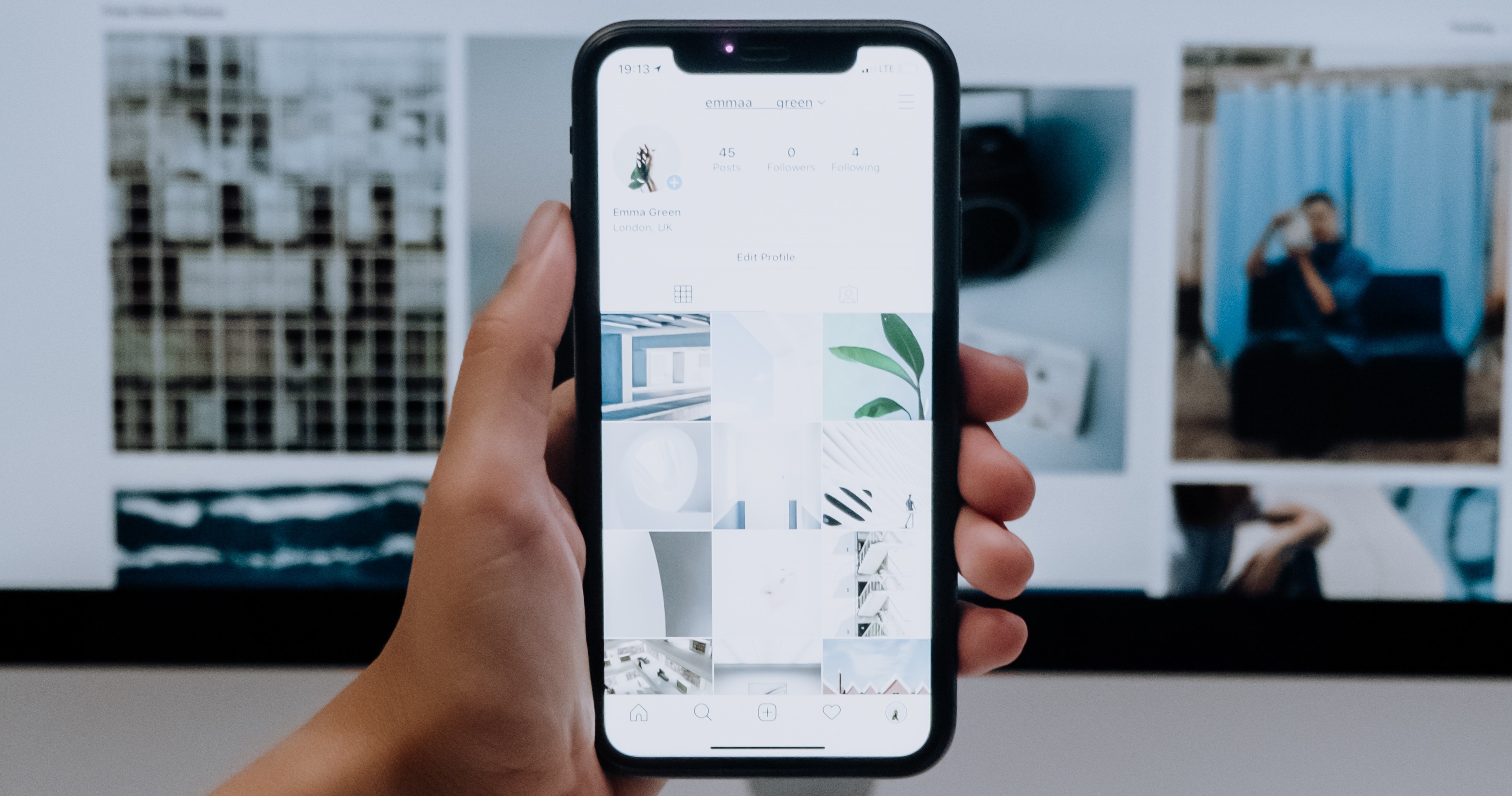 A person holds a smartphone displaying an Instagram profile in their hand, with a computer screen showing different images blurred in the background.