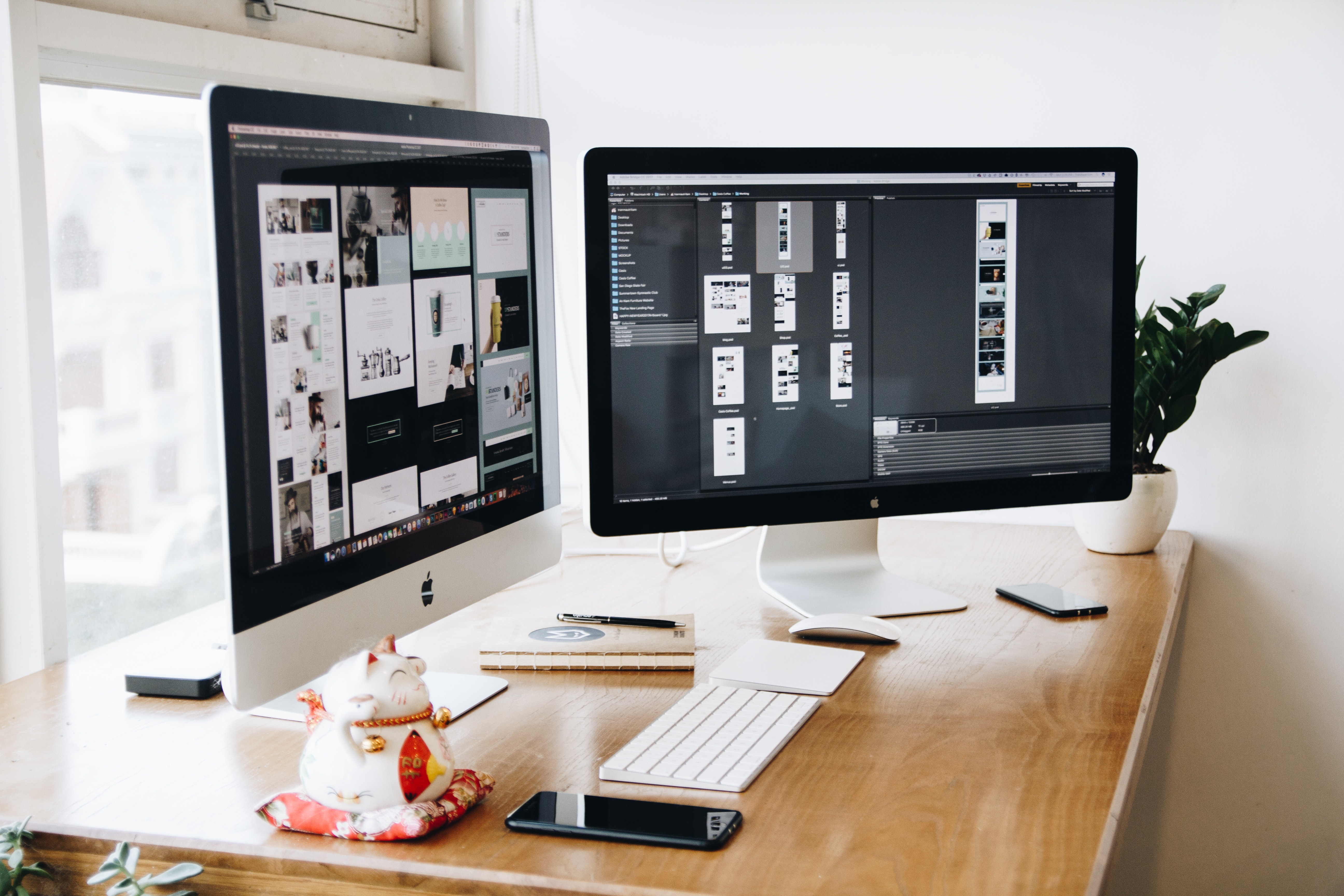 A tidy desk setup with two desktop computers displaying various design projects, along with a keyboard, mouse, smartphone, a potted plant, and a ceramic cat figurine.