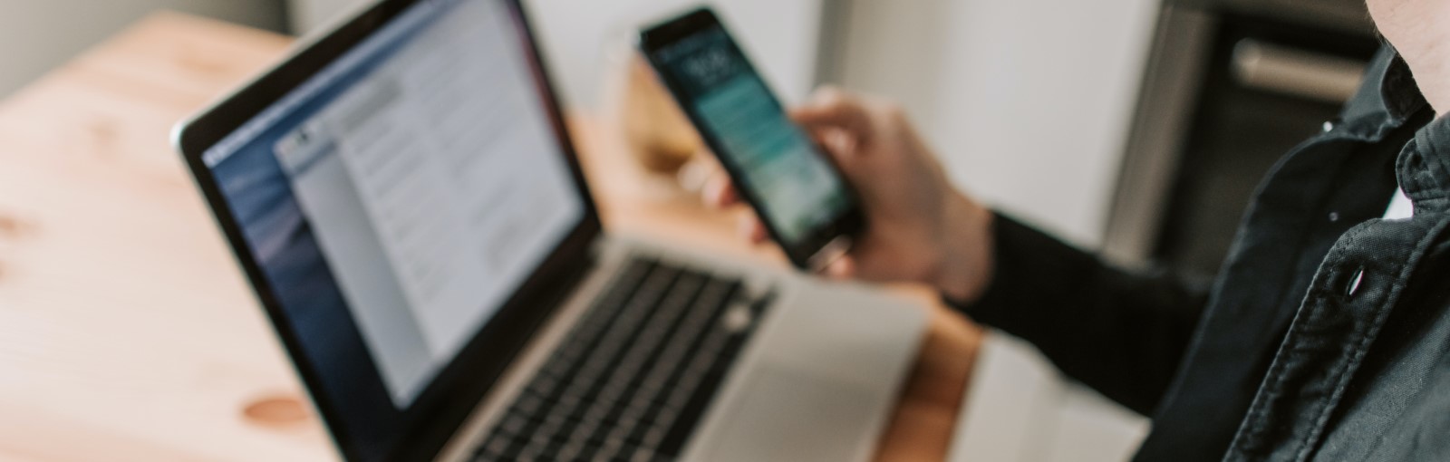 A person holds a smartphone in one hand while working on an open laptop on a wooden table.