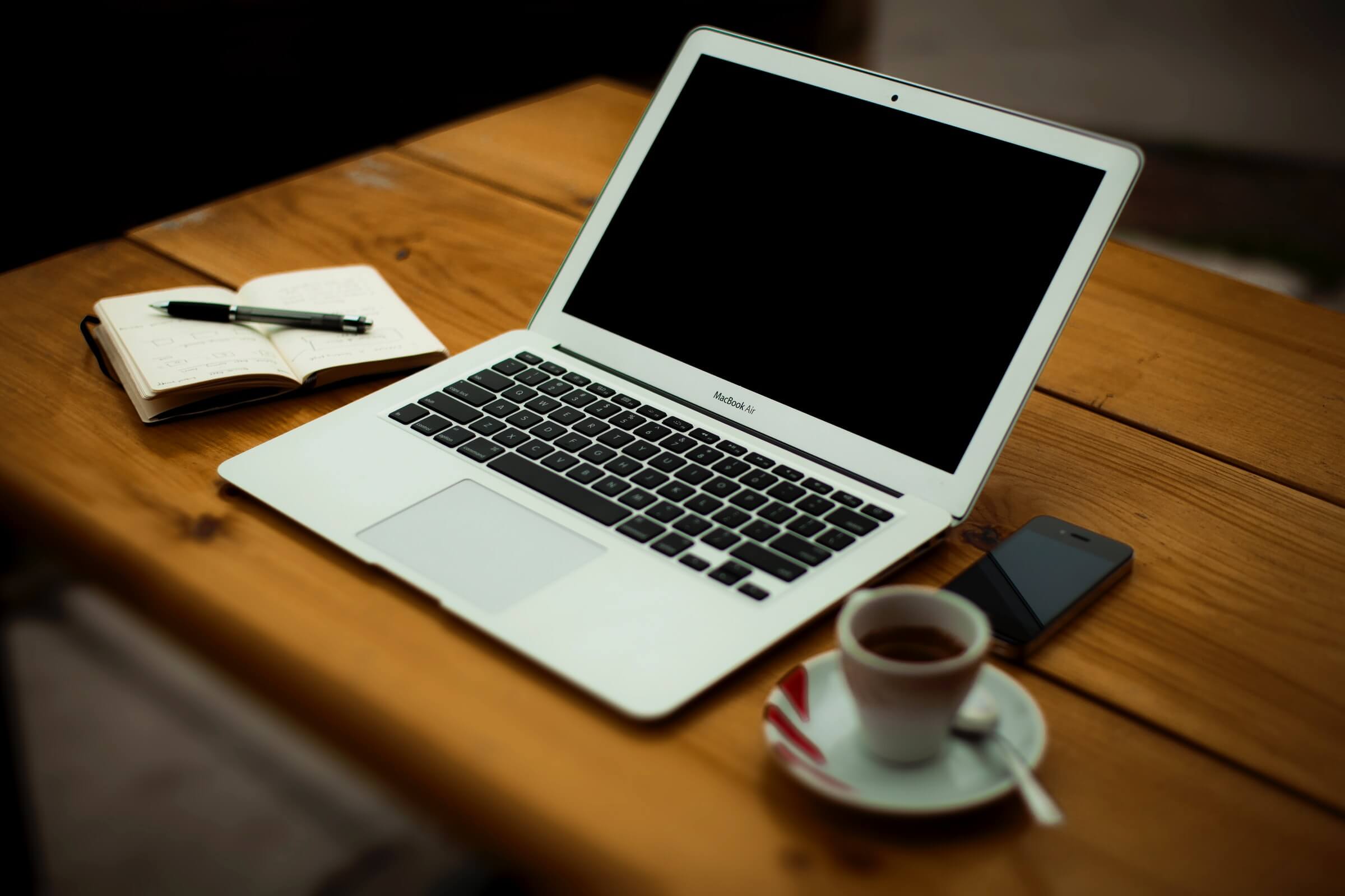 A laptop with a blank screen is on a wooden table alongside a notebook, pen, cup of coffee, and a smartphone.