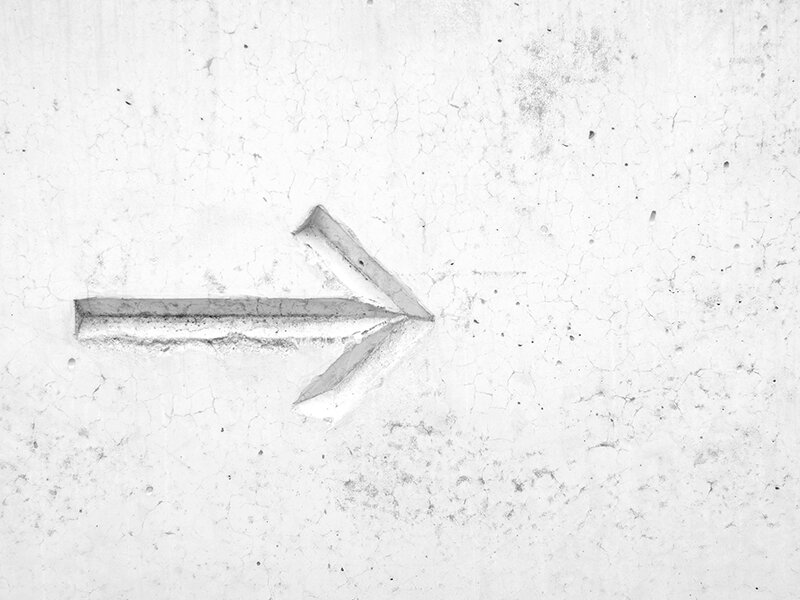 A white wall with a simple, textured arrow pointing to the right carved into its surface reminds one of navigating a new path. The wall shows signs of cracking and wear, much like the challenges in ensuring your website migration is a success.