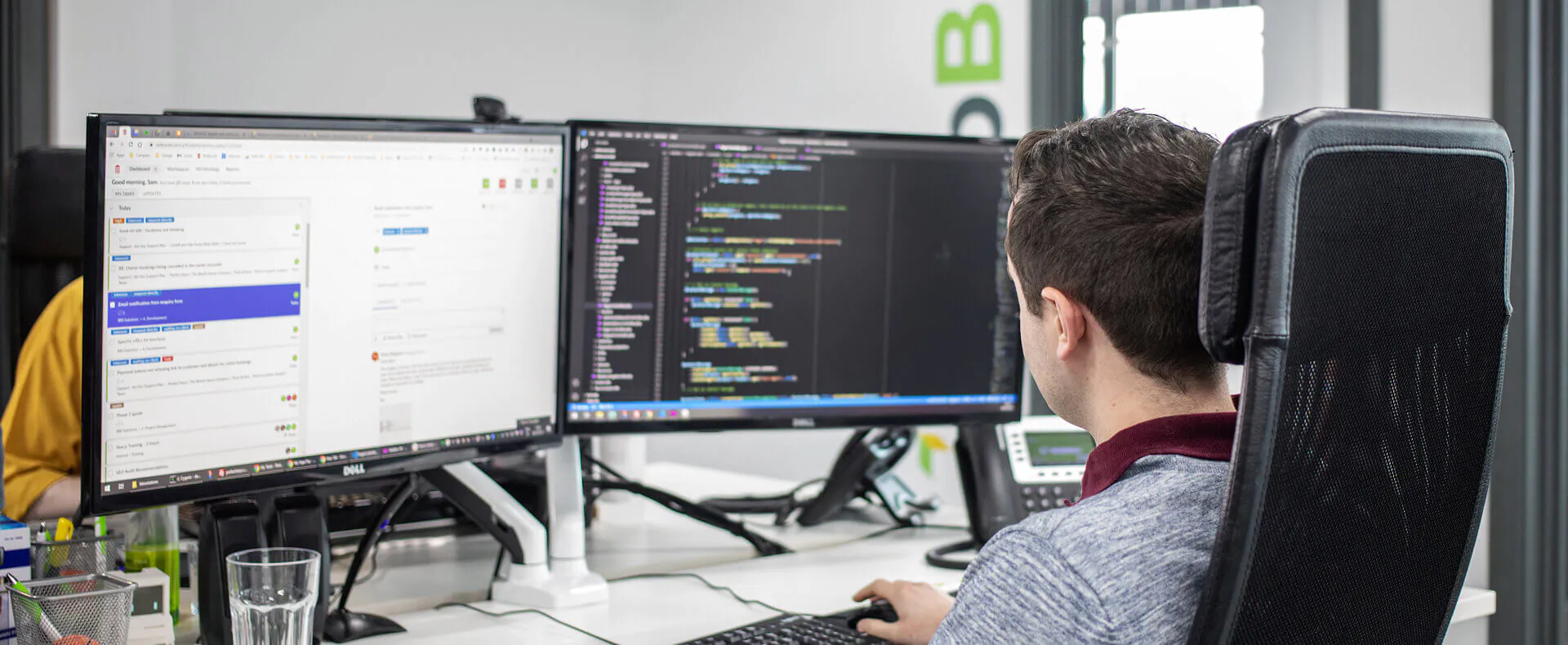 A person sits at a desk working on a dual-monitor setup. The left screen shows a page with texts and icons, while the right screen displays a code editor with multiple lines of code.