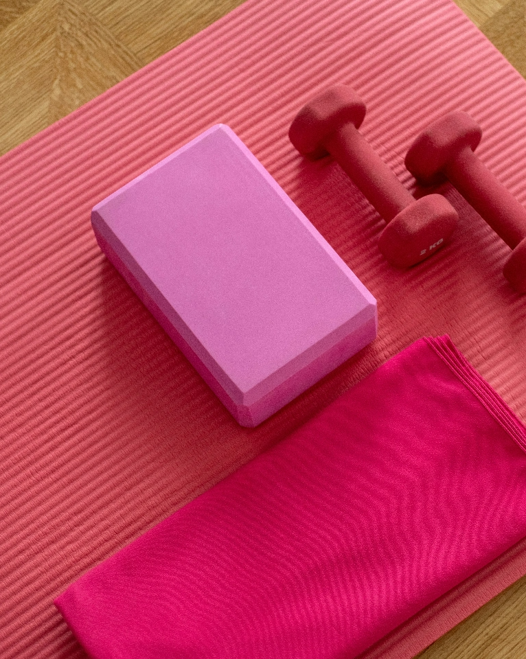 A pink yoga mat with a yoga block, two red dumbbells, and a red cloth laid on it.