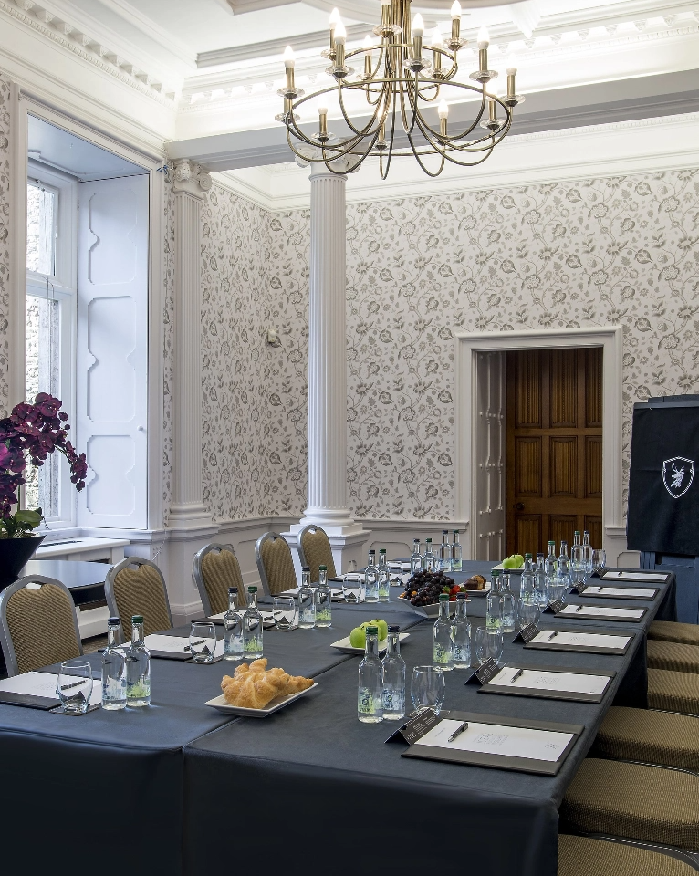A conference room features a U-shaped table setup with chairs, water bottles, notepads, pens, and a platter of pastries. The room has elegant decor with a chandelier, patterned wallpaper, and large windows.