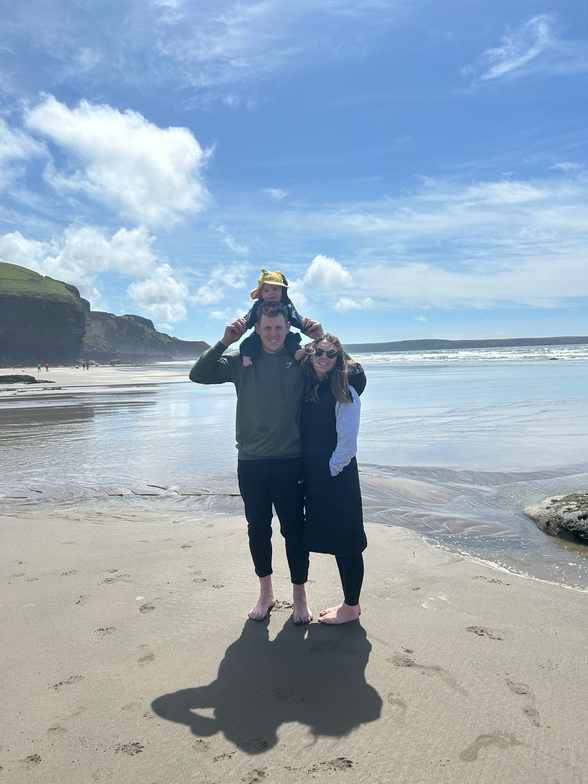 A family of three stands on a sandy beach with the ocean and cliffs in the background. The man and woman hold a child on their shoulders, and all smile at the camera under a partly cloudy sky.