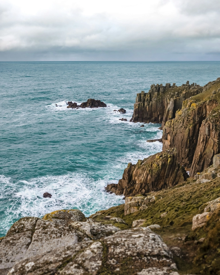 Rocky coastline with jagged cliffs overlooking rough sea waves under a cloudy sky.