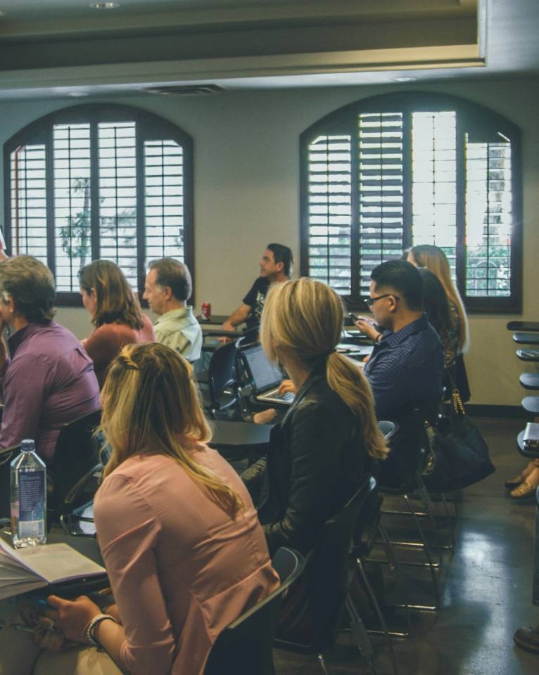 People seated in a classroom with laptops and notebooks, listening intently to a presenter at the front. The room has large windows with wooden shutters and several rows of desks with chairs.