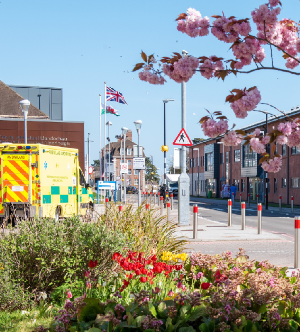 A street scene with an ambulance, blooming pink and red flowers in the foreground, and the United Kingdom flag flying on a flagpole in the background on a sunny day.