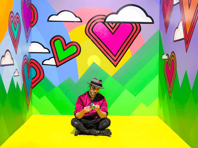A man in a pink shirt and gray hat sits on a yellow floor against a vibrant, colorful mural with hearts, clouds, and mountains.