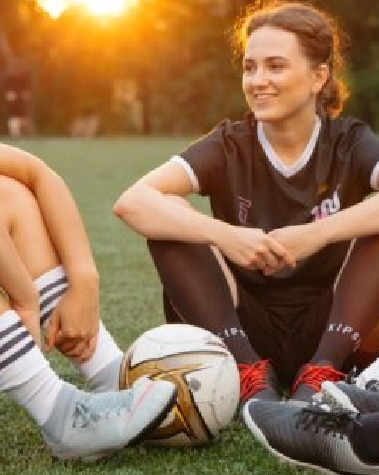 Two soccer players sit on grass with a soccer ball between them, one wearing black and the other grey, against a sunset backdrop.