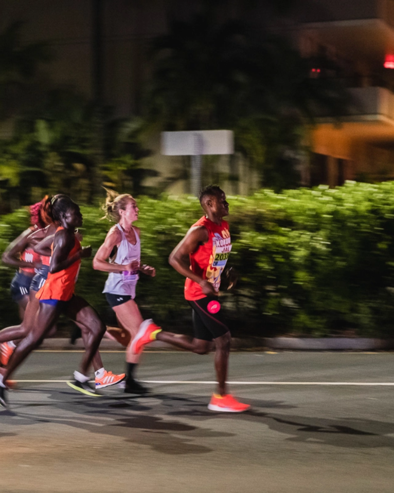 Four runners are competing in a race at night, moving swiftly along a paved road, with greenery and a building in the background.