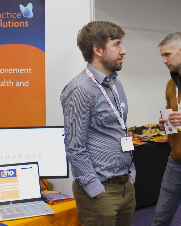 Two men are in conversation at an exhibition booth. One man is wearing a blue shirt and a lanyard, while the other is holding a booklet. A monitor displays content on a table behind them.