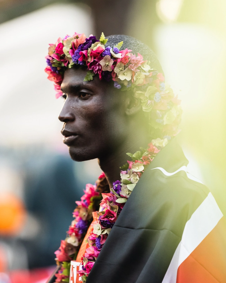 A person wearing a colorful flower crown and wrapped in a flag stands outdoors, profile view with a serious expression.
