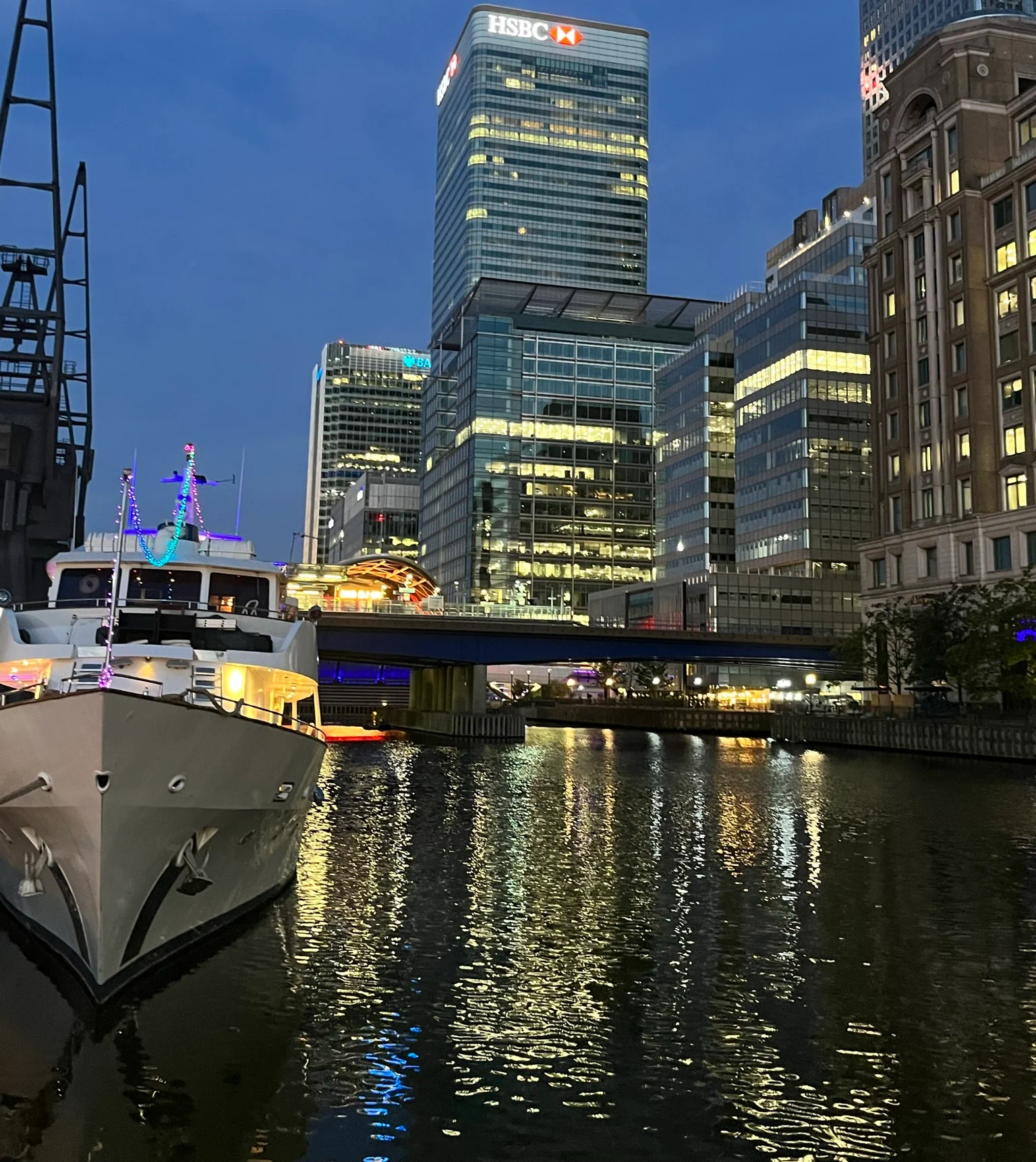 A boat moored on a waterway in a city at dusk, with illuminated high-rise office buildings and a bridge in the background.