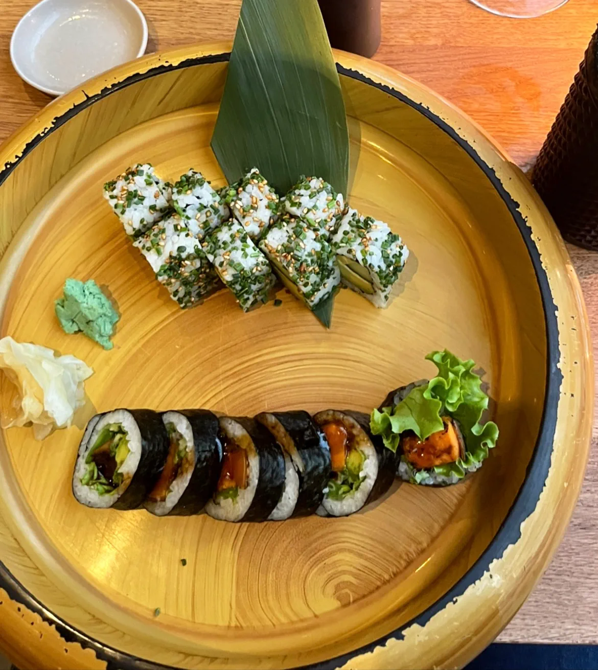 A round wooden sushi platter with two types of sushi rolls, garnished with wasabi, pickled ginger, and a leaf.