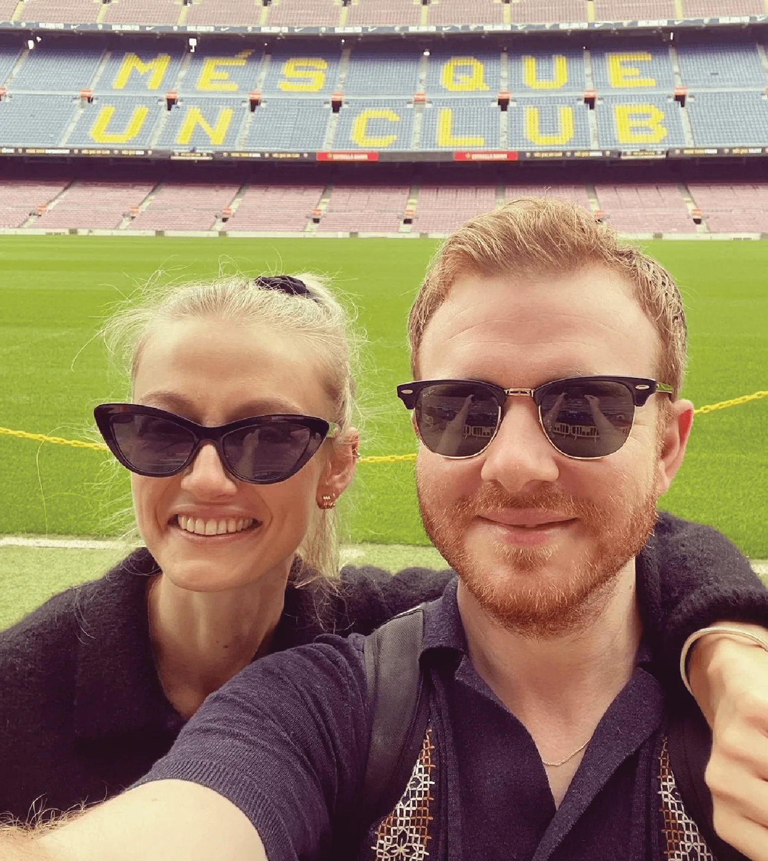 Two people wearing sunglasses take a selfie in front of a large football stadium with "MES QUE UN CLUB" written on the seats.