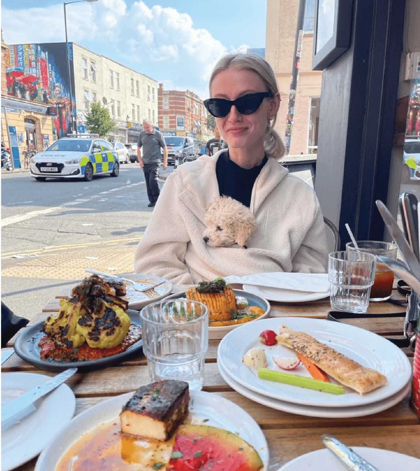 A woman in sunglasses sits at an outdoor restaurant table with various plates of food. She has a small dog in her lap and police vehicles are seen in the background on the street.