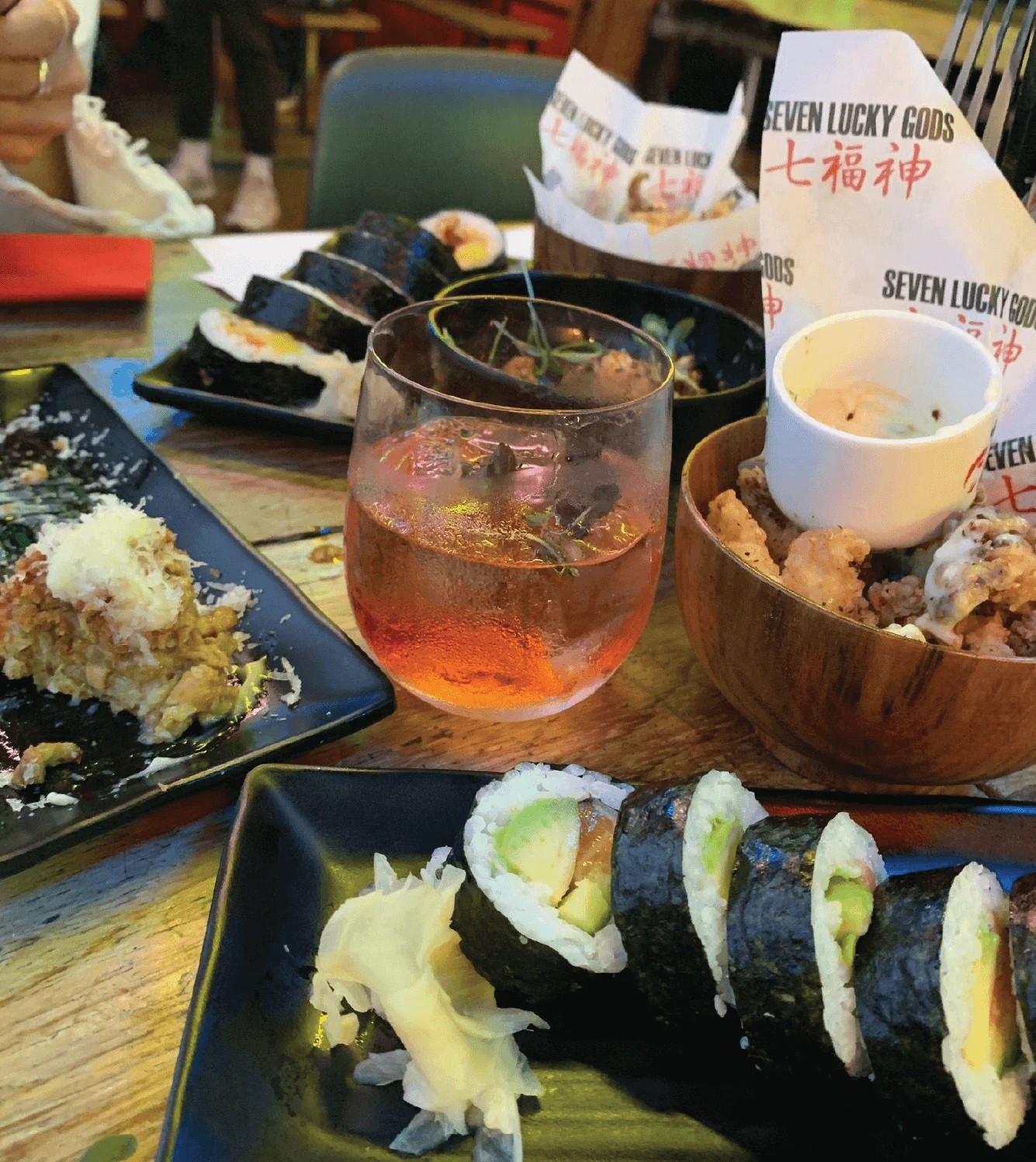 A table with sushi rolls, a cocktail, fried dishes, and ginger slices, labeled with "Seven Lucky Gods.