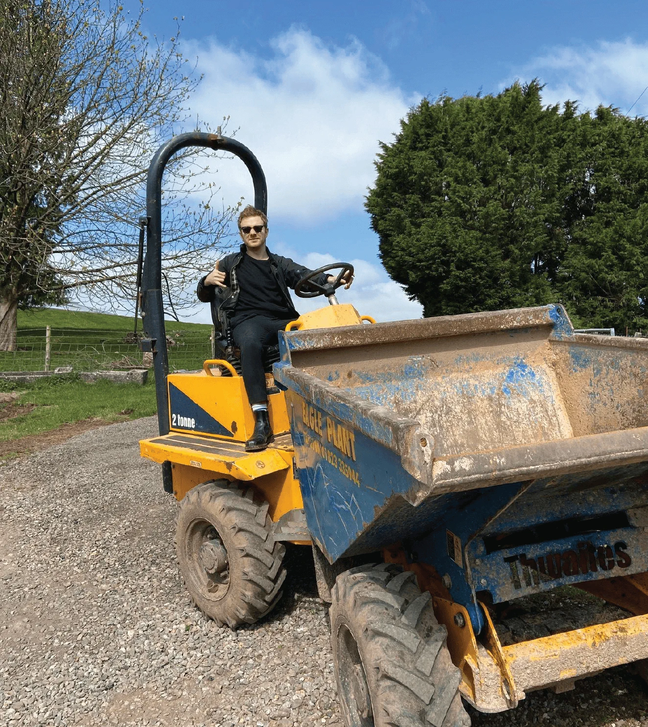 A person in dark clothing and sunglasses is sitting on a yellow and blue construction vehicle on a gravel path with trees and a field in the background.