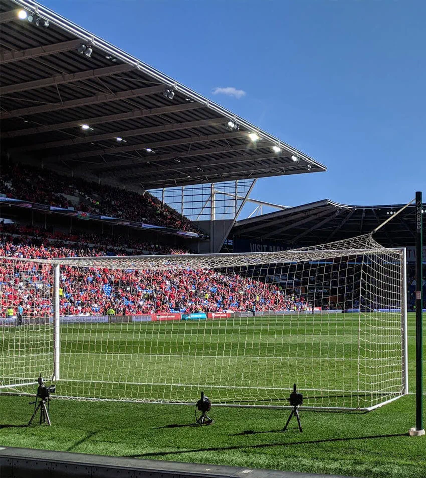 View of a soccer stadium from behind a goalpost; the stands are filled with spectators, and the sky is clear.