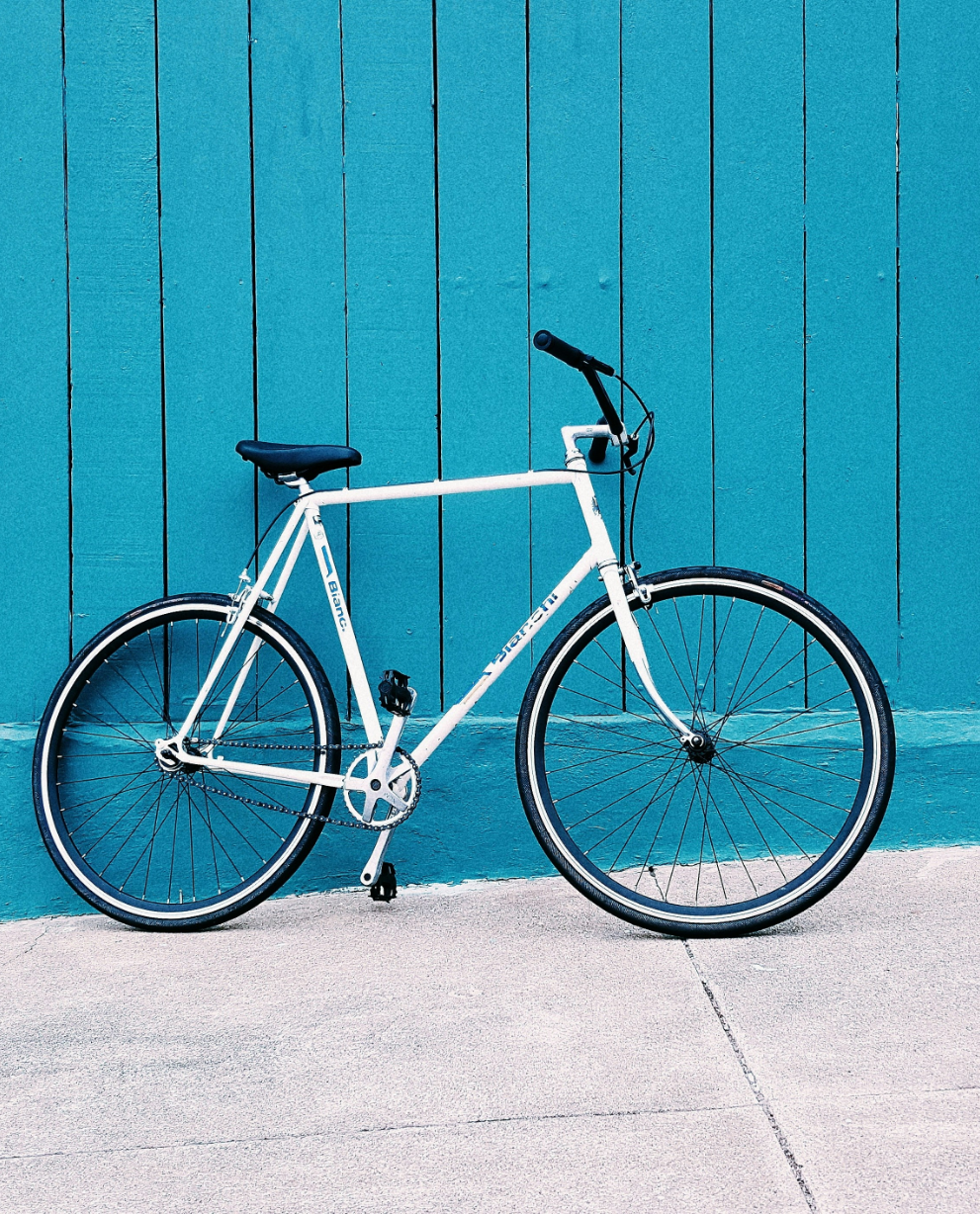 A white bicycle with black handlebars and seat is parked against a blue wooden paneled wall on a concrete sidewalk.