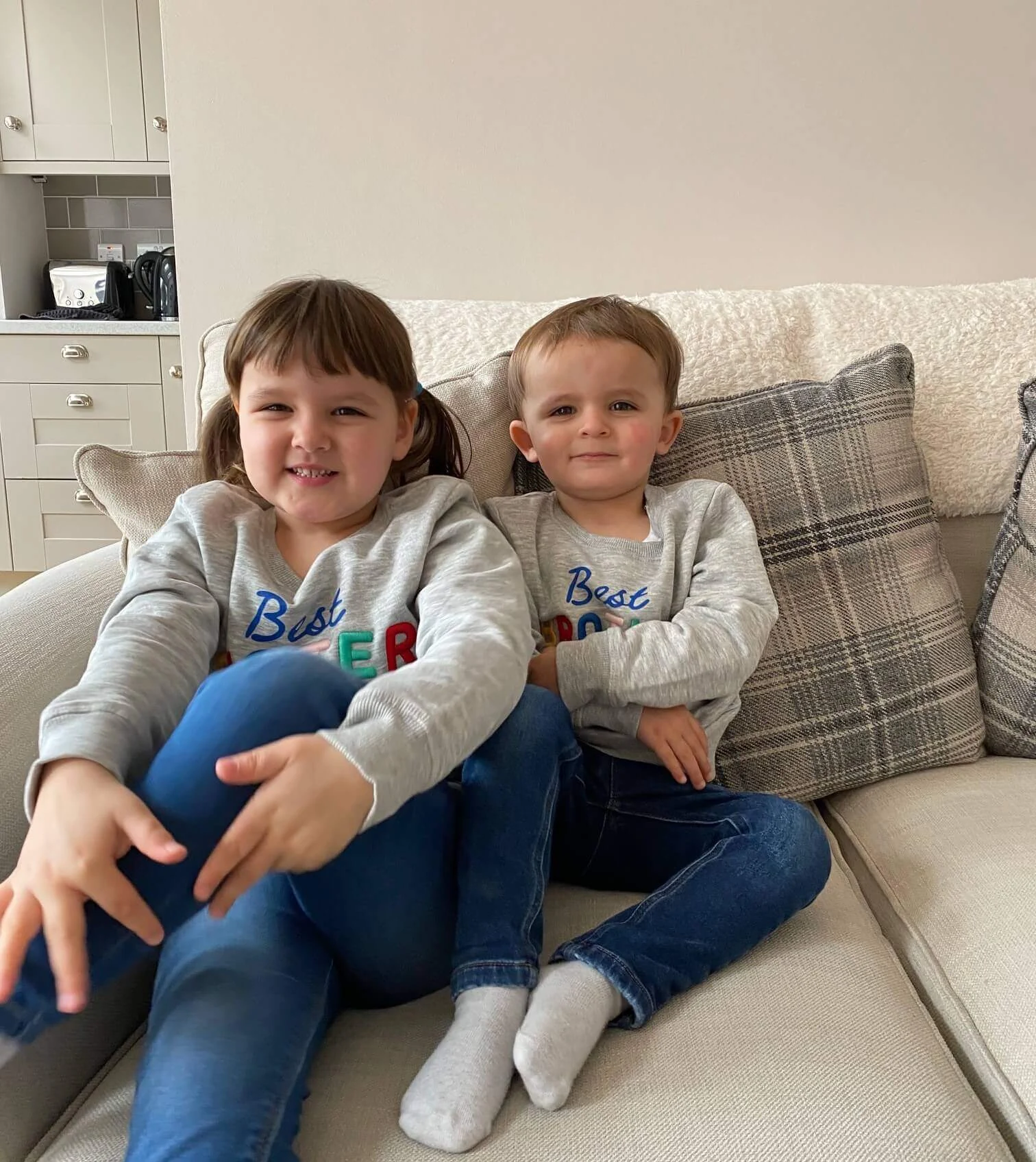 Two children sit on a beige sofa, smiling. Both are dressed in grey sweaters and blue jeans. A kitchen is visible in the background.