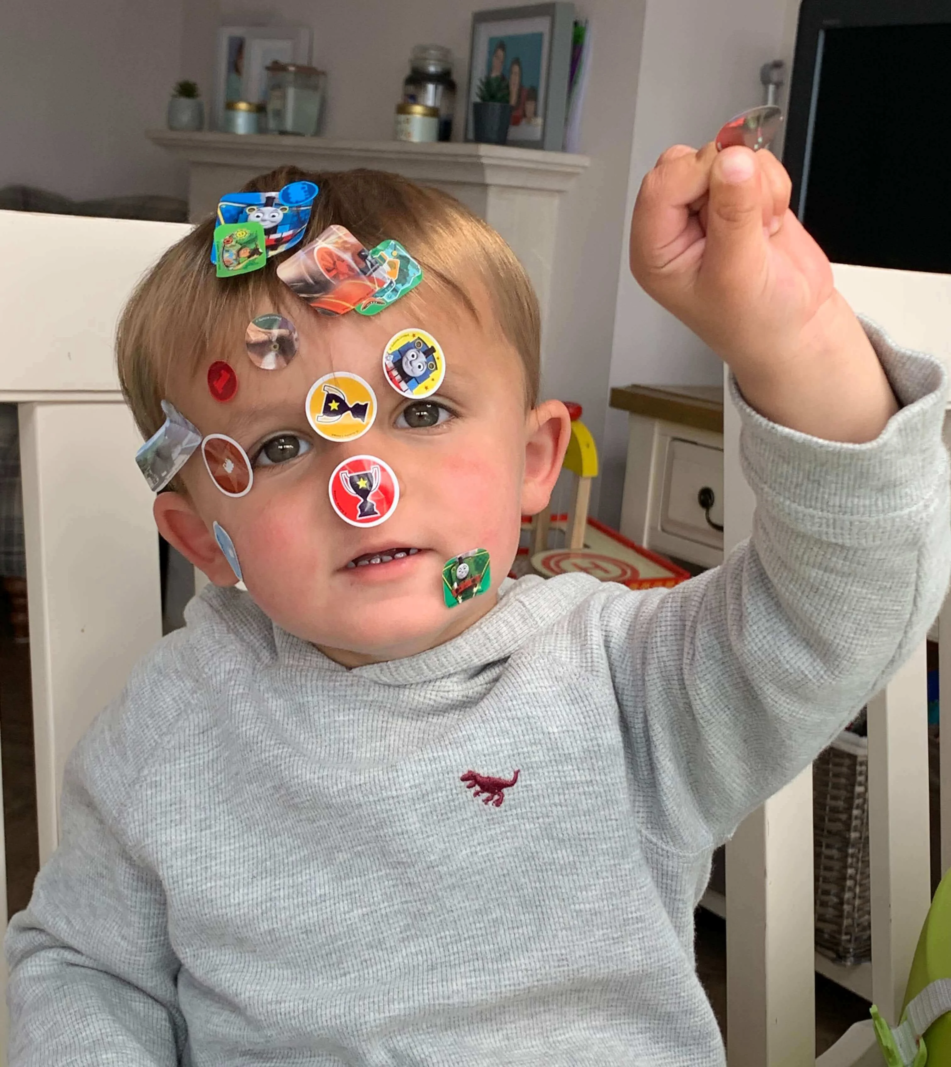 A young child with several colorful stickers on their face raises one arm while sitting on a chair.