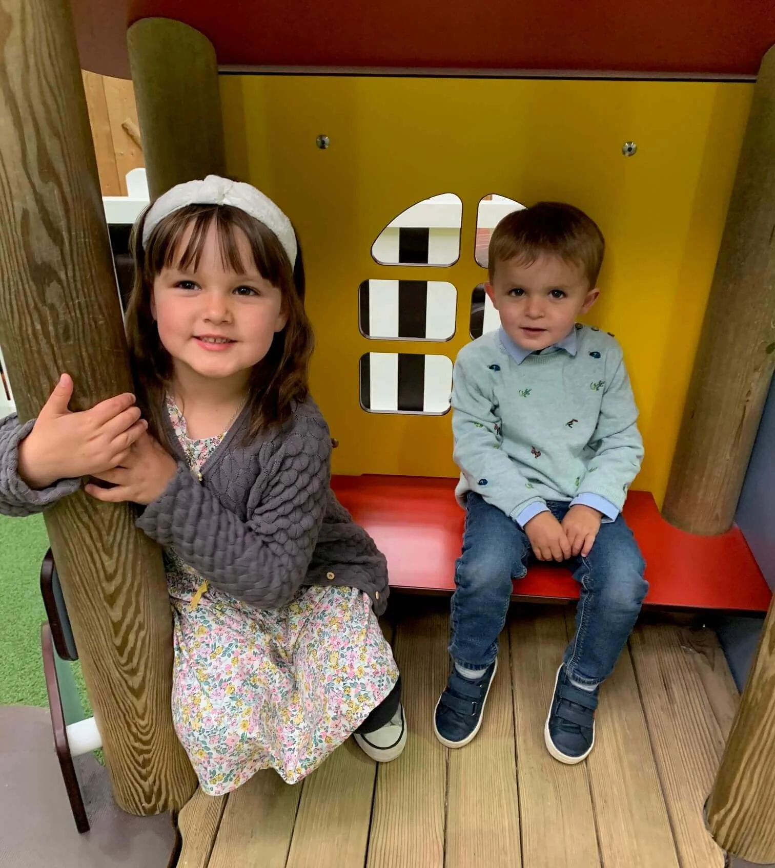 Two children are sitting in a colorful playhouse. The girl on the left is holding a wooden pole and smiling, while the boy on the right sits with his hands on his lap, looking at the camera.
