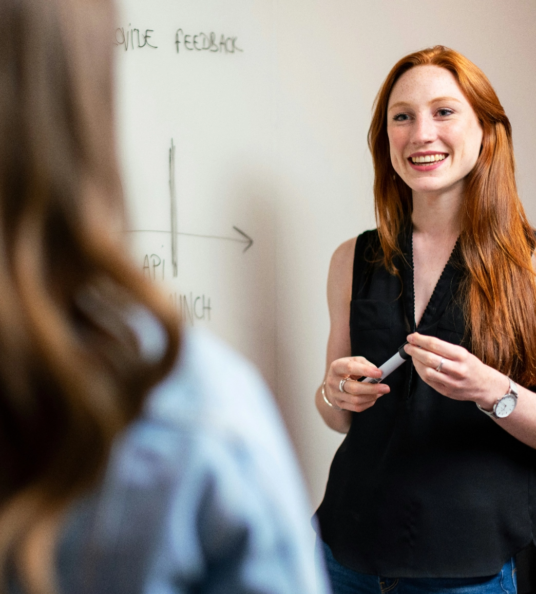 A person with red hair is smiling while holding a whiteboard marker and engaging with another person in a casual setting. A graph is partially visible on the whiteboard behind them.