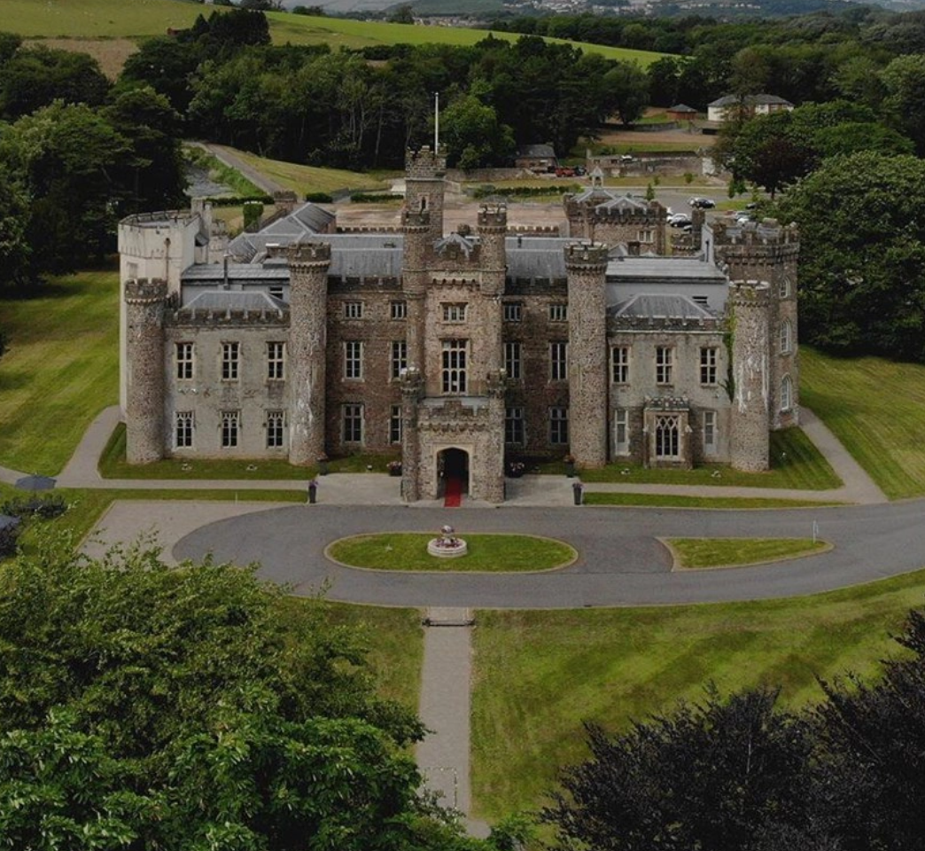 Aerial view of a large historic castle with multiple towers and a central courtyard surrounded by lush greenery and paths.