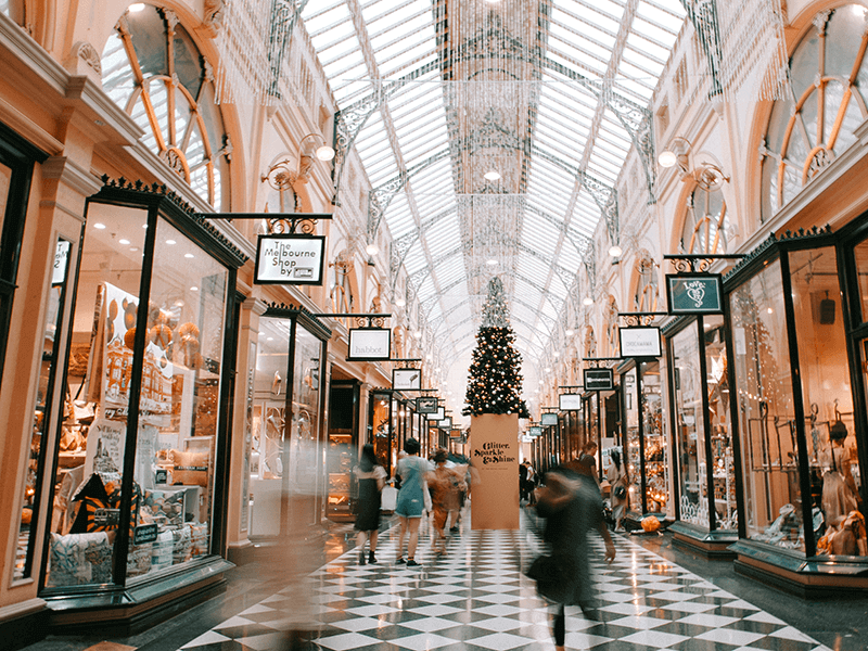 People walking through a shopping mall with a high glass ceiling, storefronts on either side, and a Christmas tree in the background.
