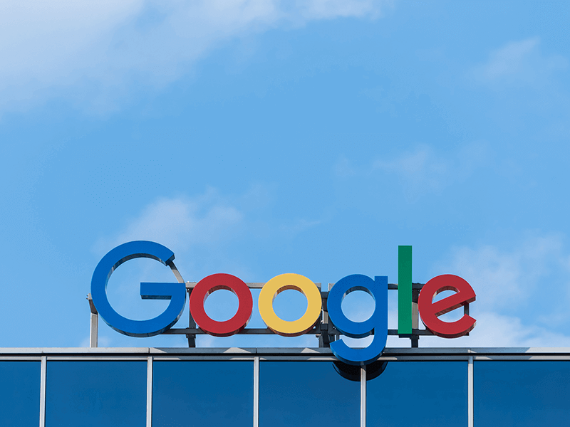 The Google logo on the top of a glass building against a clear blue sky background illustrates the tech giant's prowess. Just as their cross-channel advertising thrives seamlessly, so too does their iconic presence in our daily lives.