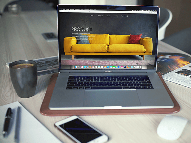 A laptop displaying a website featuring a yellow couch is on a desk, alongside a smartphone, a notebook, a magazine, and a cup of coffee.
