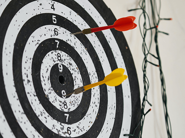 Two darts are stuck in a dartboard. One is in the number 6 region, and the other is in the number 8 region. String lights are hanging near the board.