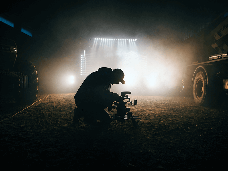 Silhouetted person in a cap kneels while operating a camera in a foggy, dimly lit environment with bright lights and large vehicles in the background, embodying the spirit of "New Year New You(Tube): The importance of video in 2020.