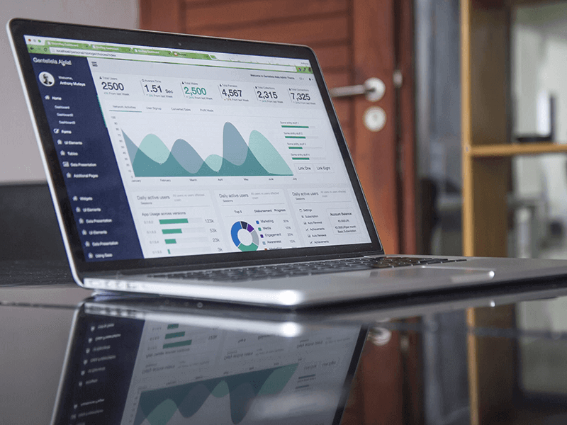 A laptop on a desk displays a data analytics dashboard with graphs and charts, showcasing 7 ways to increase your website conversion rate — fast! The glossy surface reflects the screen's vibrant visuals, while a wooden door and shelving unit form the backdrop.