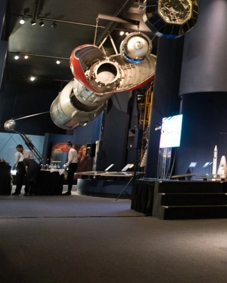 A space exhibit featuring a large, suspended spacecraft with visitors observing below in a dimly lit museum setting.