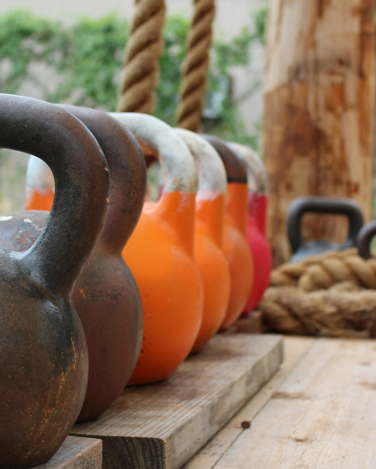 A row of colorful kettlebells arranged on a wooden platform with thick ropes in the background.