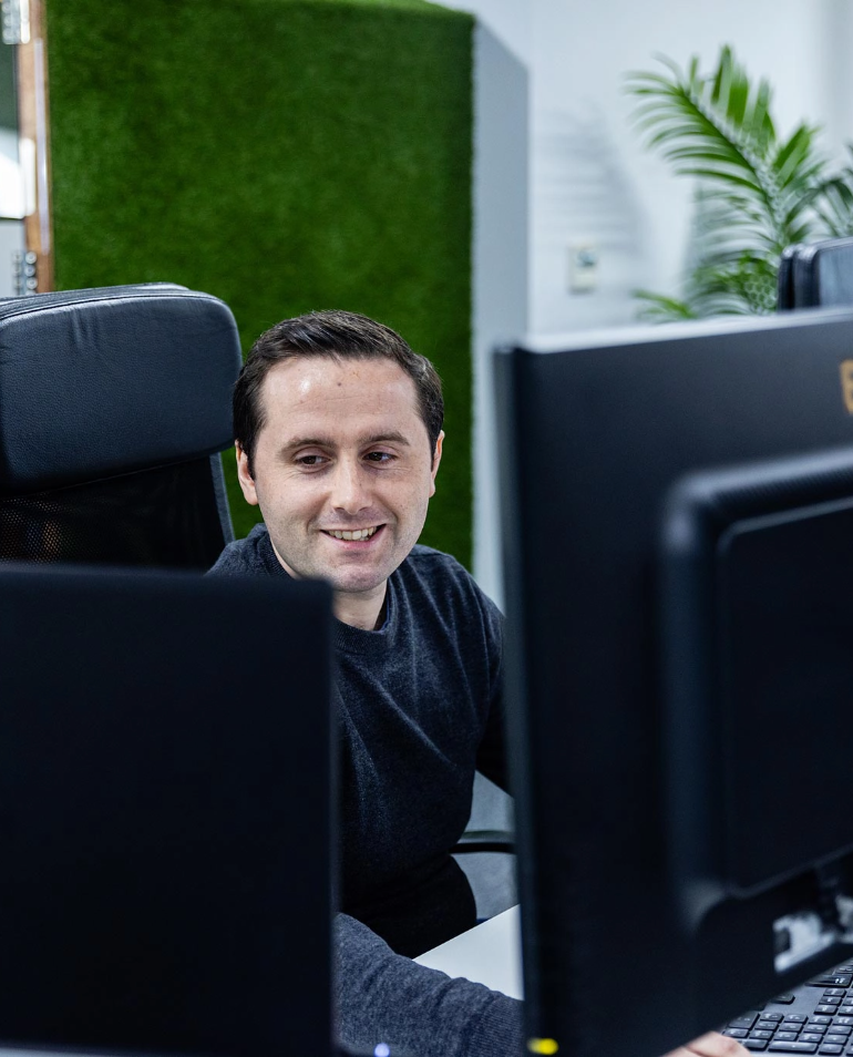 A man seated at a desk smiles while working on a computer in an office setting with a plant and green wall in the background.