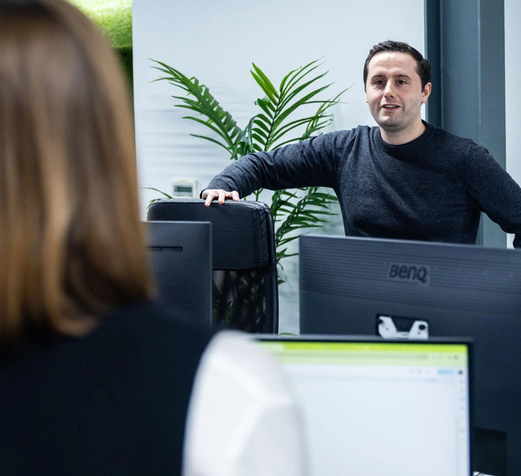 A man leaning on a chair speaks to a woman in an office with computer monitors and a plant in the background.
