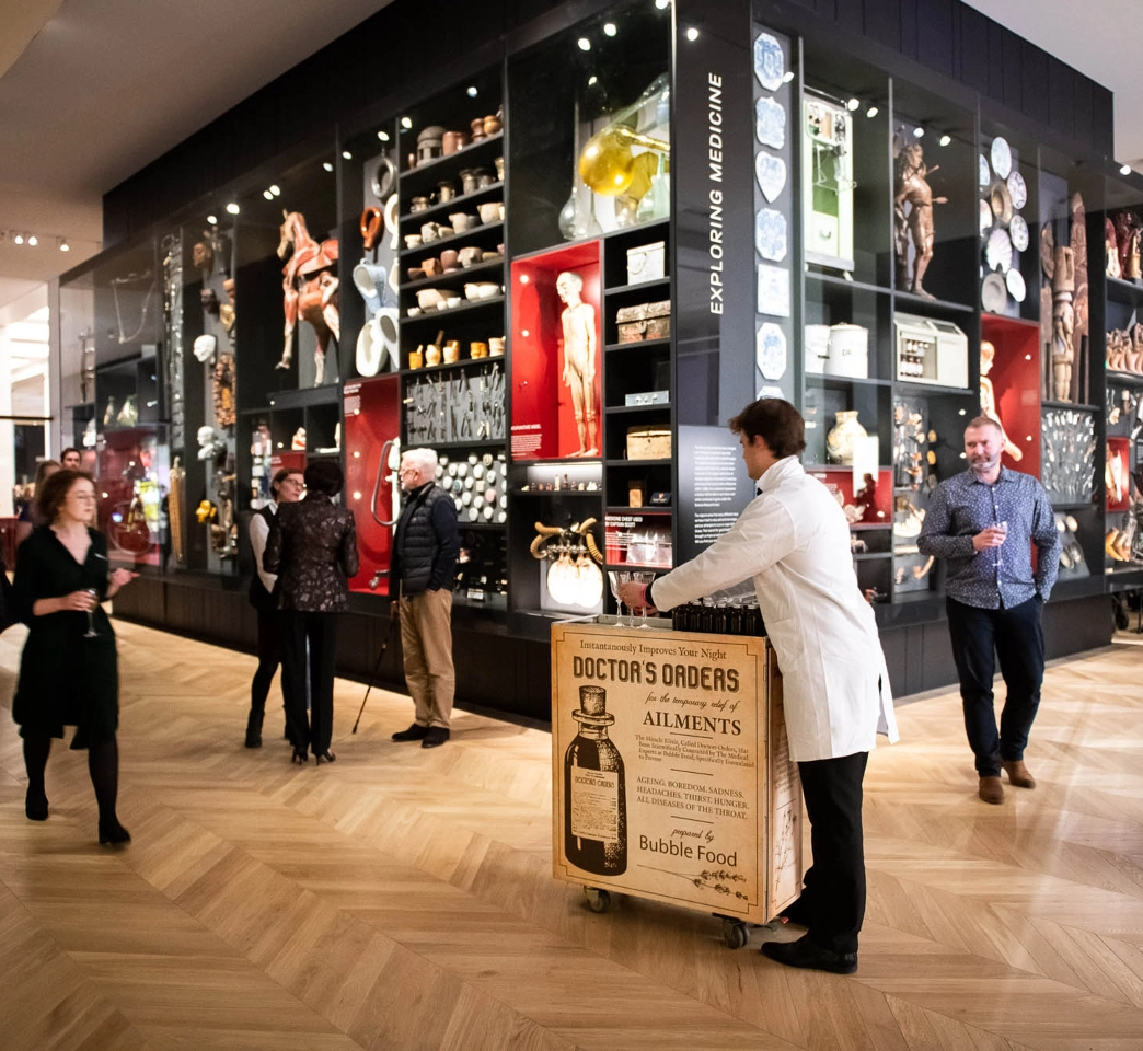 A person in a white coat pushes a cart labeled "Doctor's Orders" in a museum with glass display cases showcasing medical artifacts. Several people observe the exhibits and interact in the background.