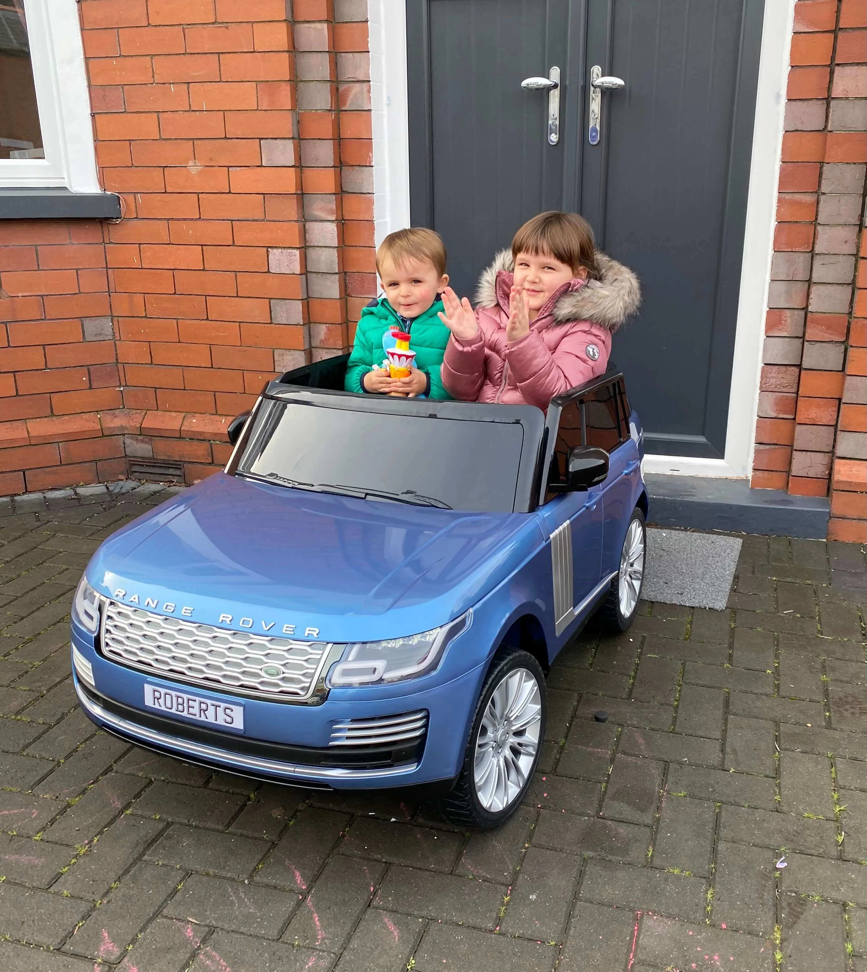 Two children are sitting in a blue toy Range Rover with a "ROBERTS" license plate, parked on a brick driveway in front of a door. The older child is clapping while the younger one holds a toy.