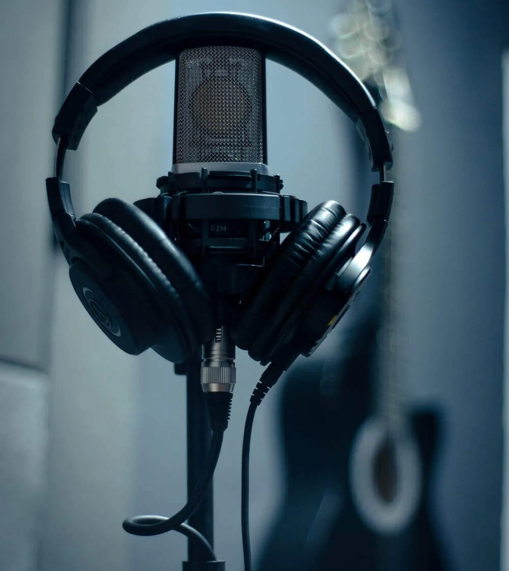 Close-up of a microphone with a pair of headphones draped over it, placed on a stand. In the blurred background, a guitar is partially visible.