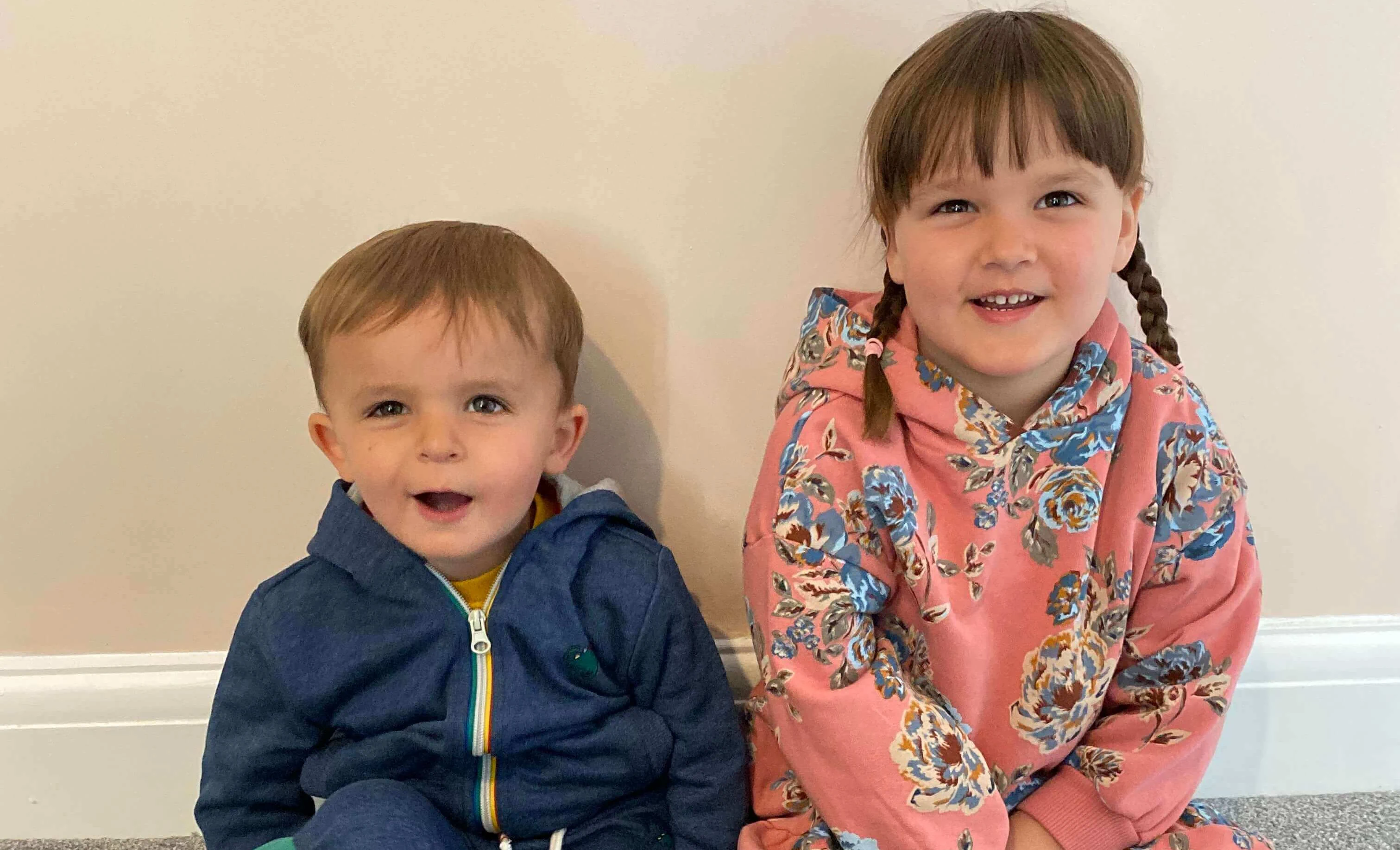 Two young children sitting on the floor against a beige wall, a boy on the left wearing a blue hoodie and a girl on the right wearing a pink floral hoodie. Both are smiling.