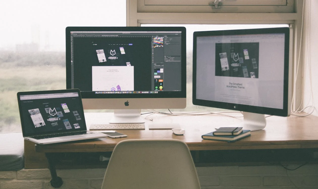 A workspace with a laptop and two desktop monitors on a wooden desk, displaying various web pages. A chair, a closed notebook, a smartphone, and a mouse are also on the desk.