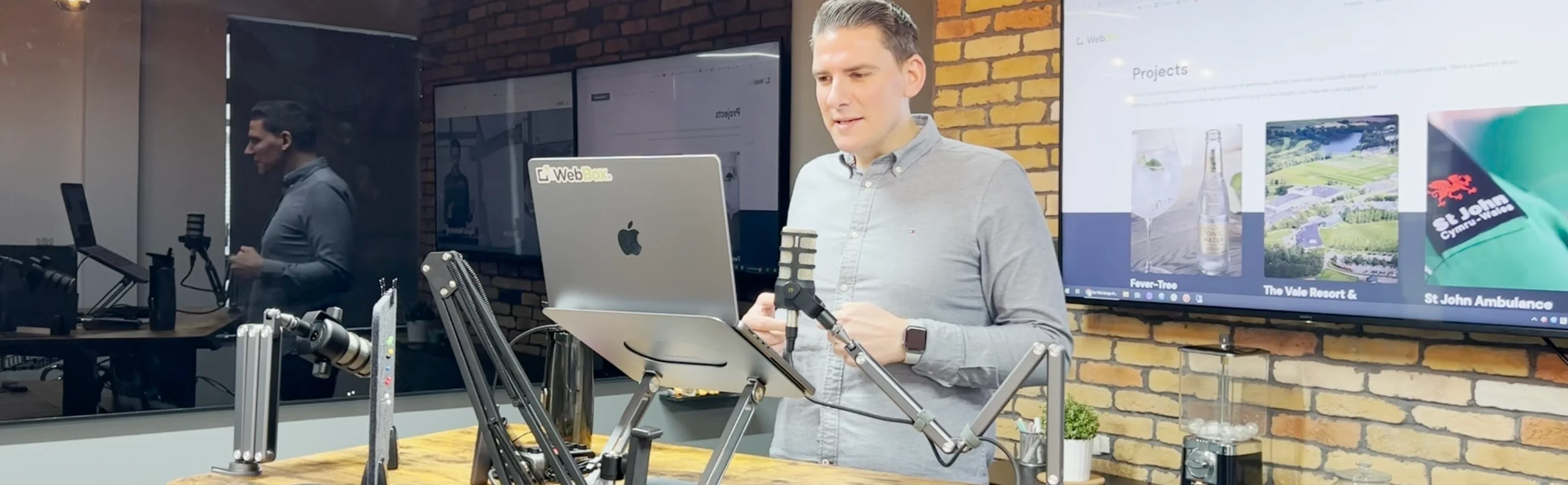 A man stands at a desk with a laptop, microphone, and other equipment in a modern office setting, preparing for an online presentation or broadcast.